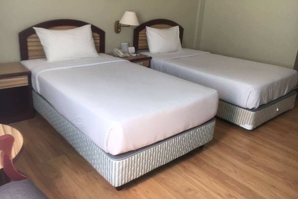 A hotel room with two single beds, wooden headboards, a bedside table with a lamp, and a small painting on the wall.