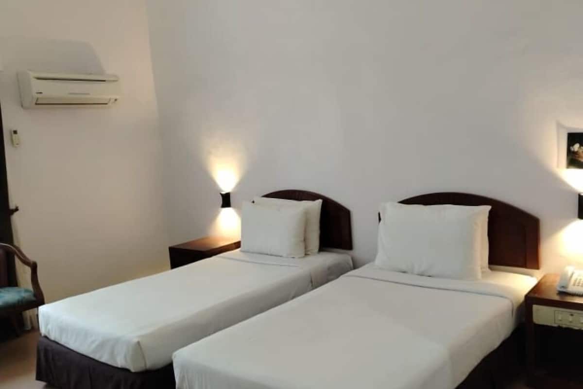 Habitación de hotel con dos camas individuales, una mesita de noche con lámpara, aire acondicionado de pared y una silla.
