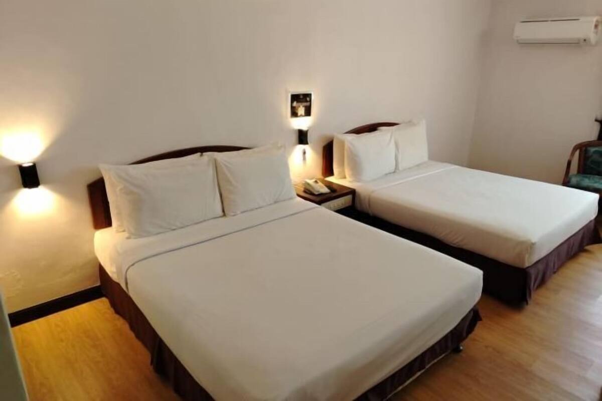 Habitación de hotel con dos camas individuales, una mesita de noche con lámpara, una pequeña mesa y un equipo de aire acondicionado en la pared.