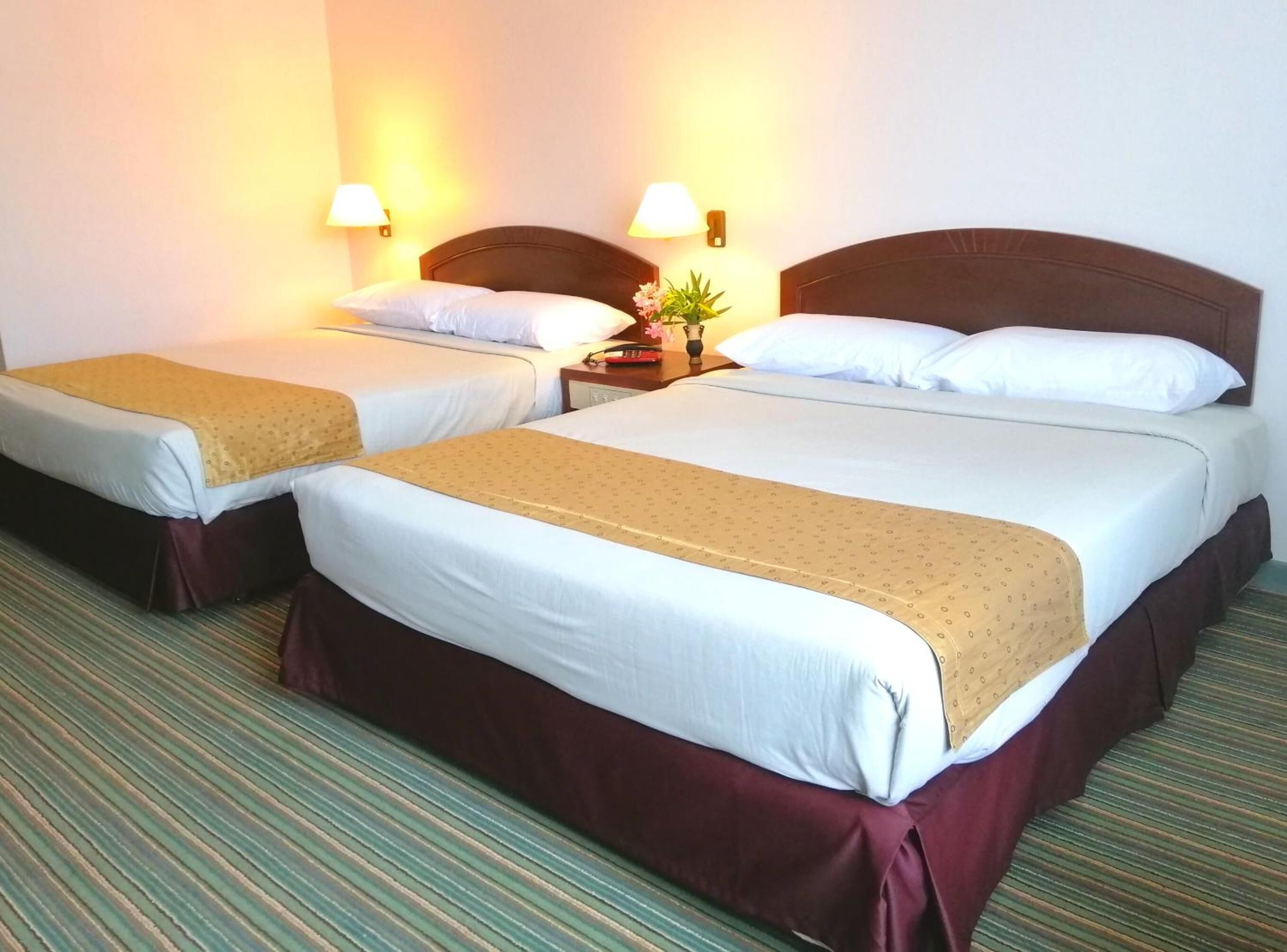 A hotel room with two beds, each with a brown headboard, white linens, and a yellow patterned throw. There are bedside lamps and a small table with a vase of flowers.