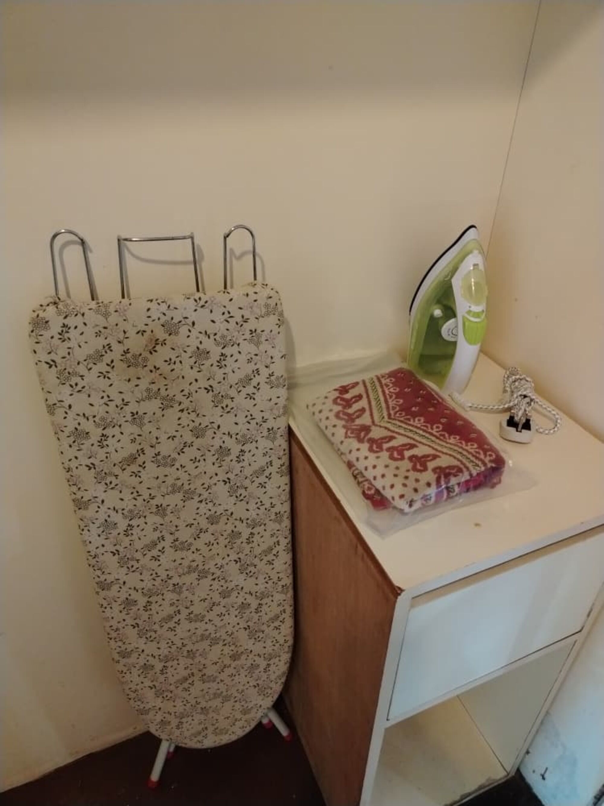 A small room with a wooden cabinet, a white ironing board with an iron, and a patterned curtain.