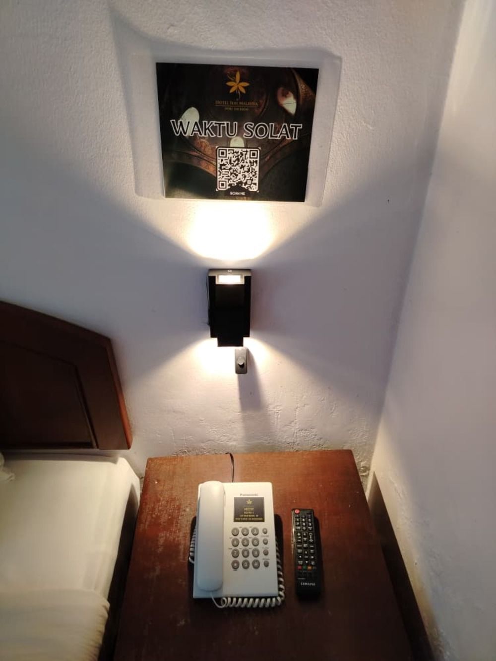 Una habitación de hotel con una mesita de noche que tiene un teléfono fijo y un control remoto. Encima de la mesita, una lámpara de pared con un letrero que dice 'WAKTU SOLAT' y un código QR.