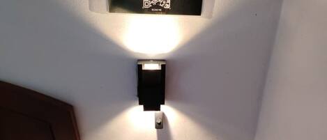 Una habitación de hotel con una mesita de noche que tiene un teléfono fijo y un control remoto. Encima de la mesita, una lámpara de pared con un letrero que dice 'WAKTU SOLAT' y un código QR.
