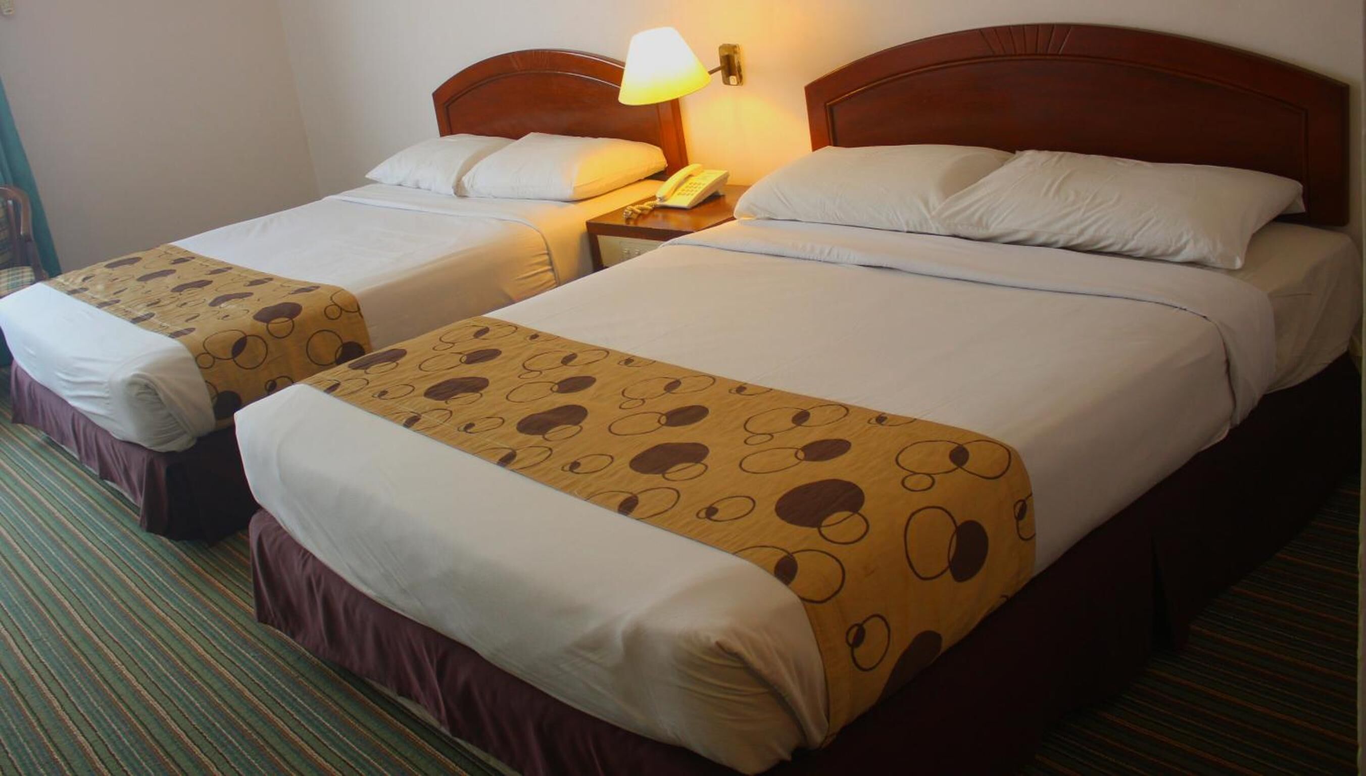 Twee eenpersoonsbedden met houten hoofdeinden, witte lakens en een geweven plaid. Elk bed heeft een nachtkastje met een lamp en een telefoon. De kamer heeft een gestreept tapijt.