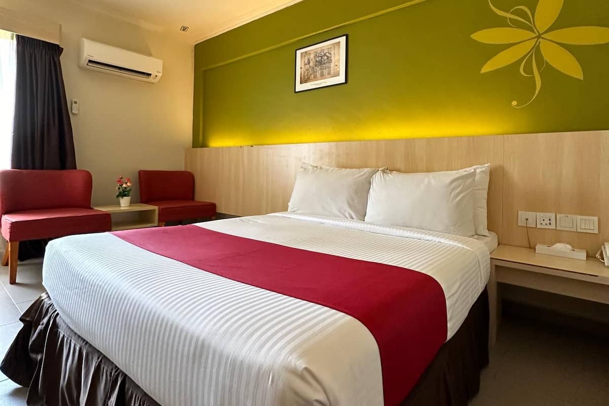 Une chambre d’hôtel avec un grand lit, une couvre-lit rouge et blanche, une tête de lit en bois, un fauteuil rouge, une petite table avec un vase de fleurs et un appareil de climatisation fixé au mur.
