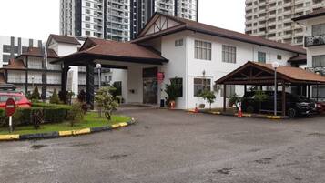 A hotel with a covered parking area, a small garden, and a building with multiple floors in the background.