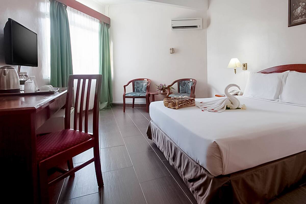 A hotel room with a bed, a desk, a chair, a TV, an air conditioner, and a window with curtains.
