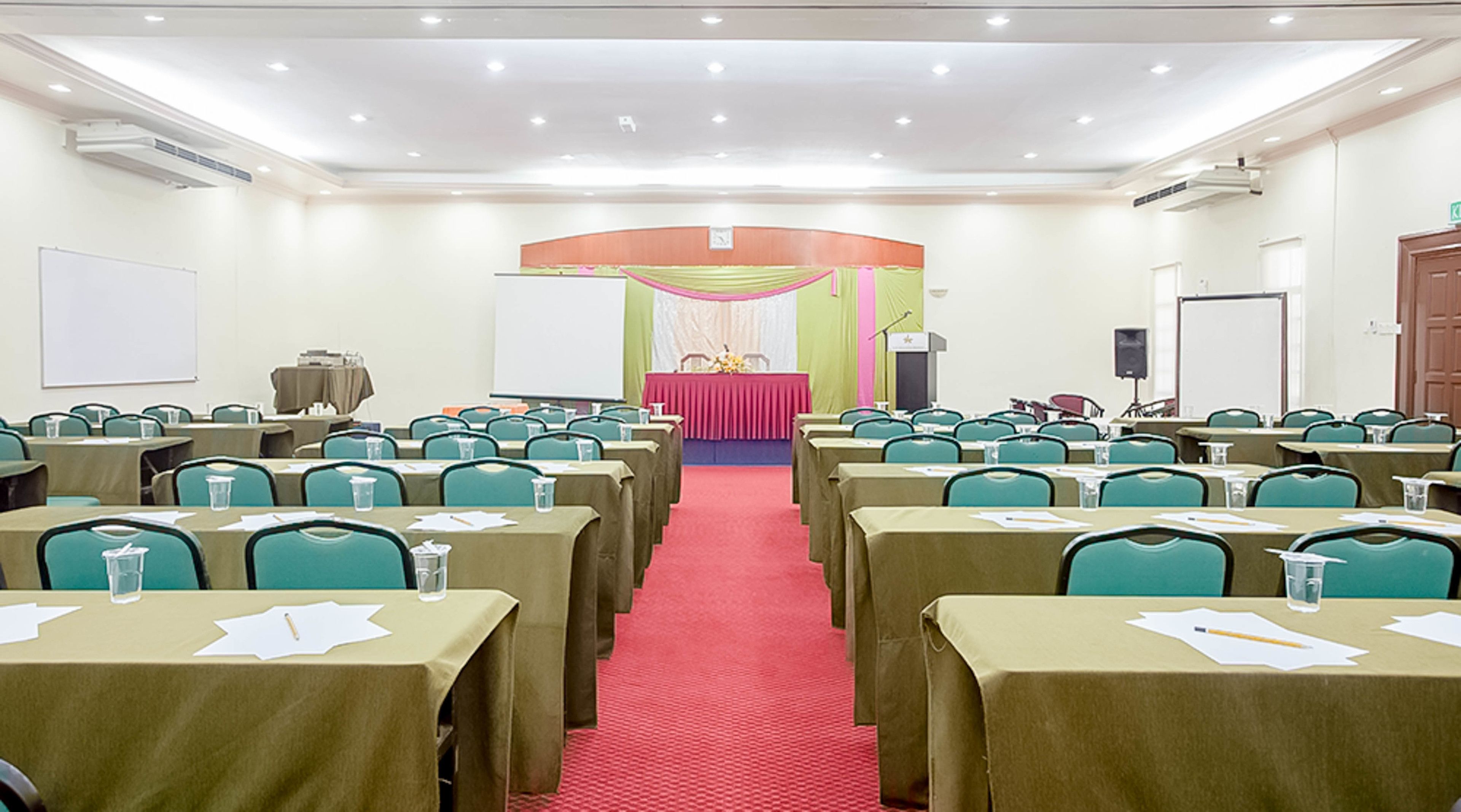 Une salle de conférence avec des rangées de tables et de chaises, un pupitre et un tableau blanc.