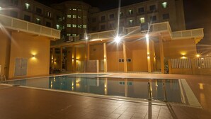 En swimmingpool med en glasvägg som avgränsning, omgiven av en byggnad med flera våningar och balkonger, samt utomhusbelysning.