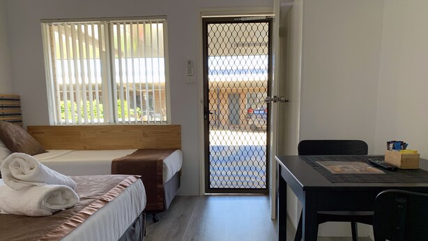 A hotel room with two beds, a wooden headboard, a desk with a chair, and a view through a door with a metal gate. - Baths Motel (Moree)