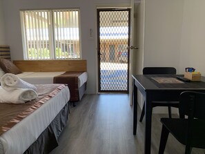 A hotel room with two beds, a wooden headboard, a desk with a chair, and a view through a door with a metal gate.