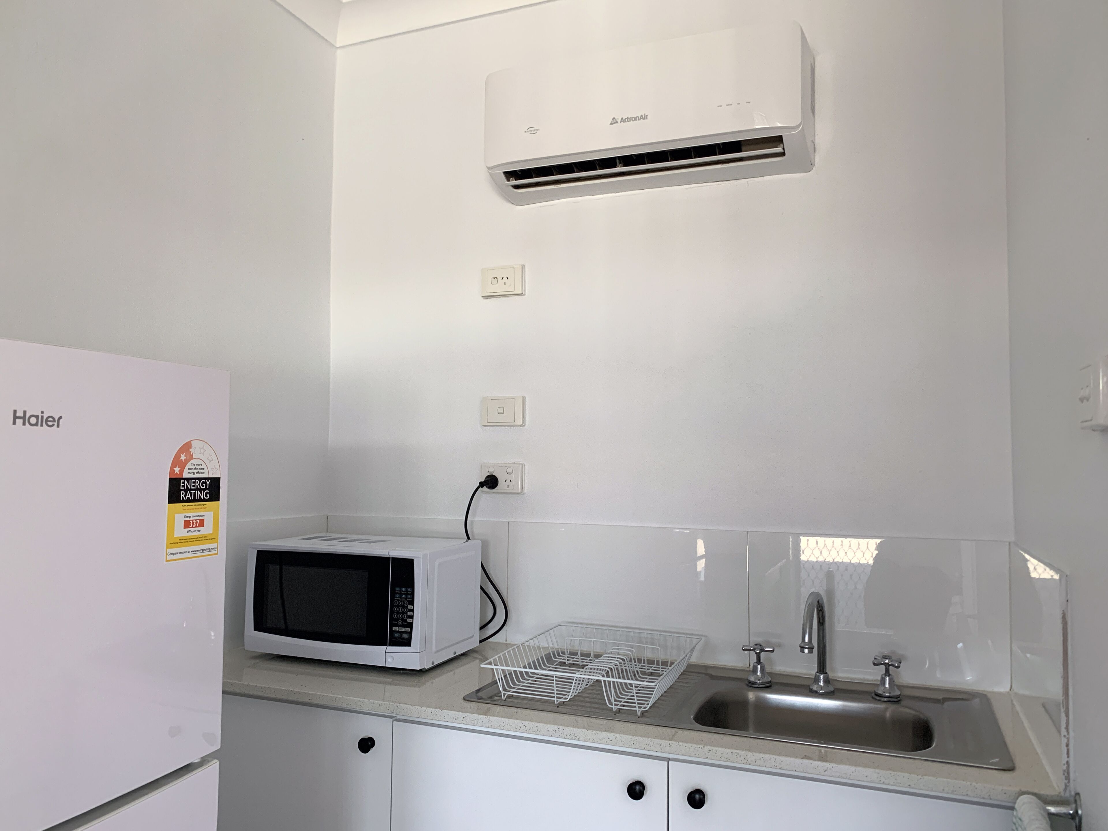 Una cocina compacta con un refrigerador blanco, un microondas, un lavamanos y un aire acondicionado.