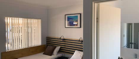 Une chambre d’hôtel avec deux lits simples, une tête de lit en bois, un tableau encadré au mur et une salle de bain visible à travers une porte ouverte.