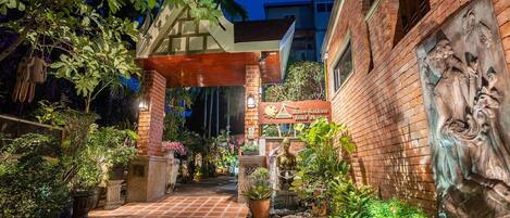 A well-lit outdoor area with a brick pathway leading to a building entrance, surrounded by lush greenery and potted plants.