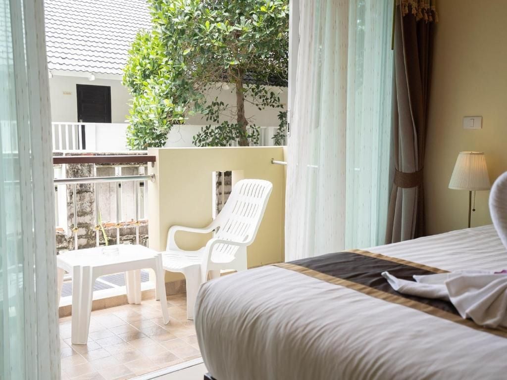 A hotel room with a bed, a table, and a chair. There is a balcony with a view of a building and a tree.