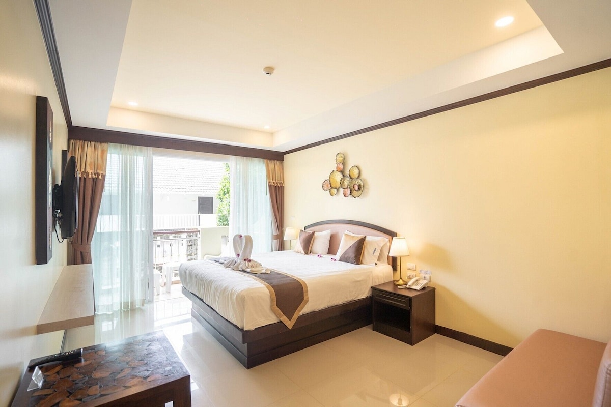A hotel room with a bed, bedside table, lamp, and a view of the outside.