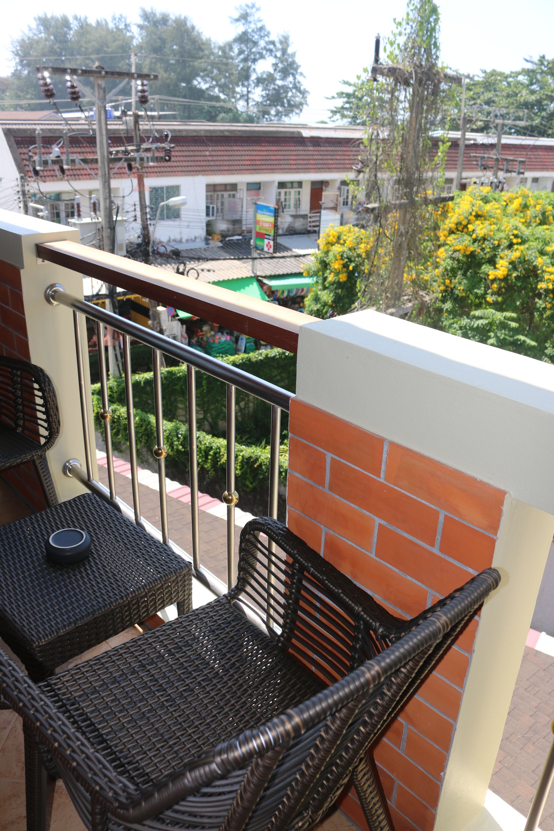 Un balcón con mobiliario de mimbre, baranda metálica y vista a una calle con edificios y vegetación.