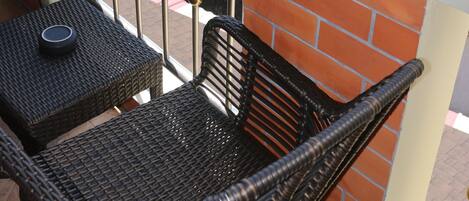 A balcony with wicker furniture, a metal railing, and a view of a street with buildings and greenery.