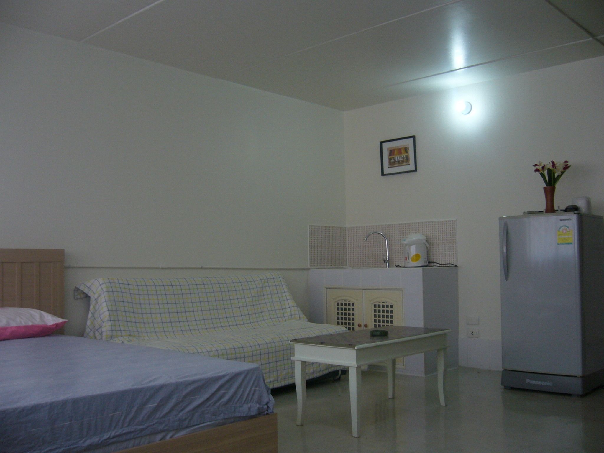 A small, sparsely furnished room with a bed, a table, a kitchenette, and a refrigerator.