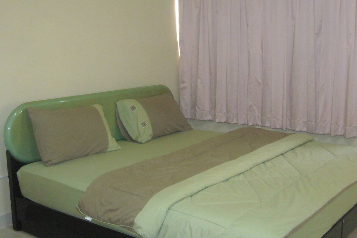 Una cama individual con cabecera verde, dos almohadas y una manta de color claro en una habitación con pared lisa y ventana con cortina blanca.
