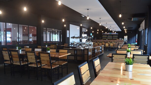 A modern cafe with wooden tables and chairs, a bar area, and large windows allowing natural light.