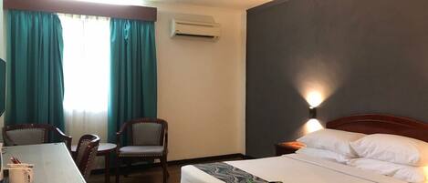 A hotel room with a bed, a desk with chairs, an air conditioner, and a window with curtains.