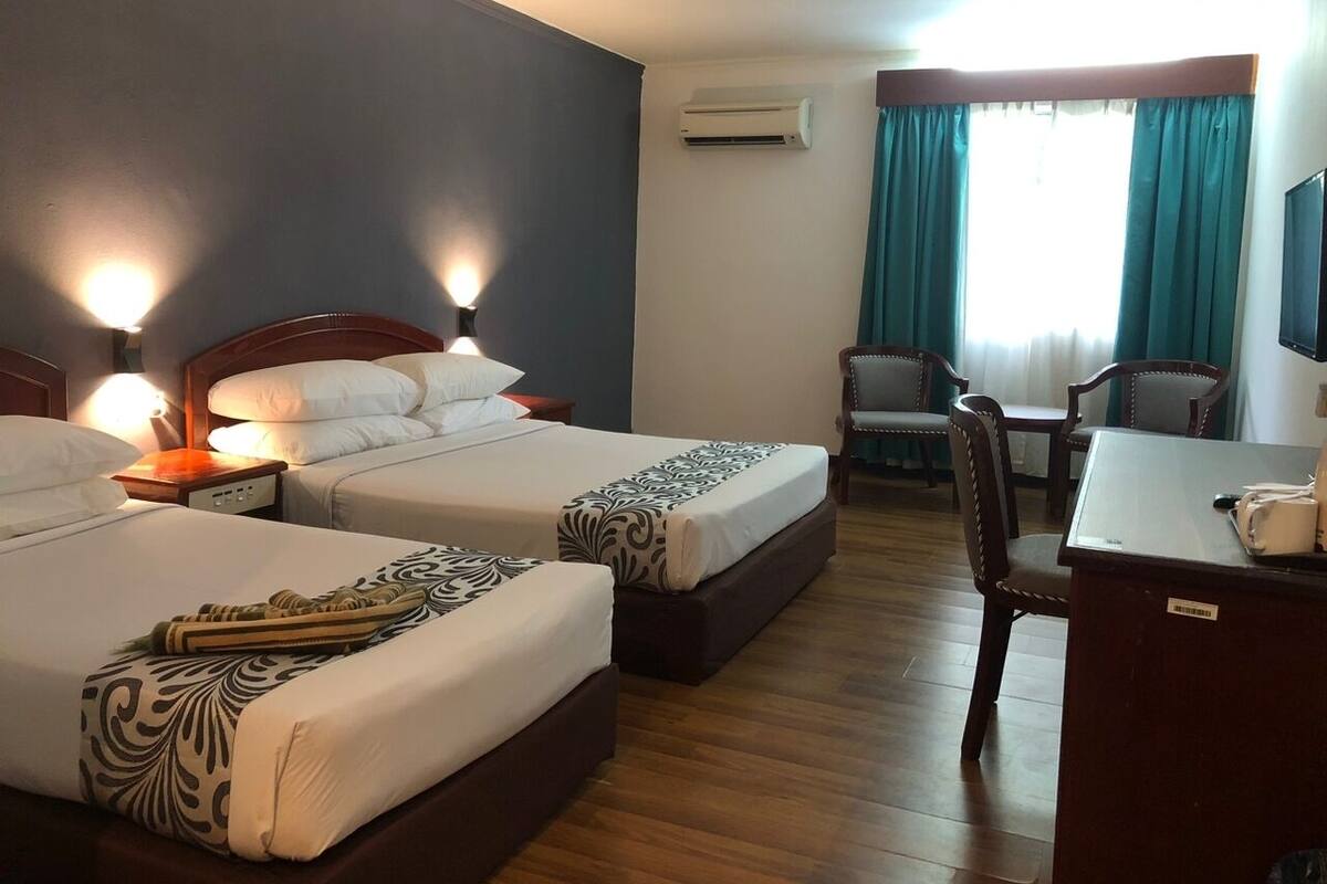 A hotel room with two beds, a desk, a chair, an air conditioning unit, and a window with curtains.