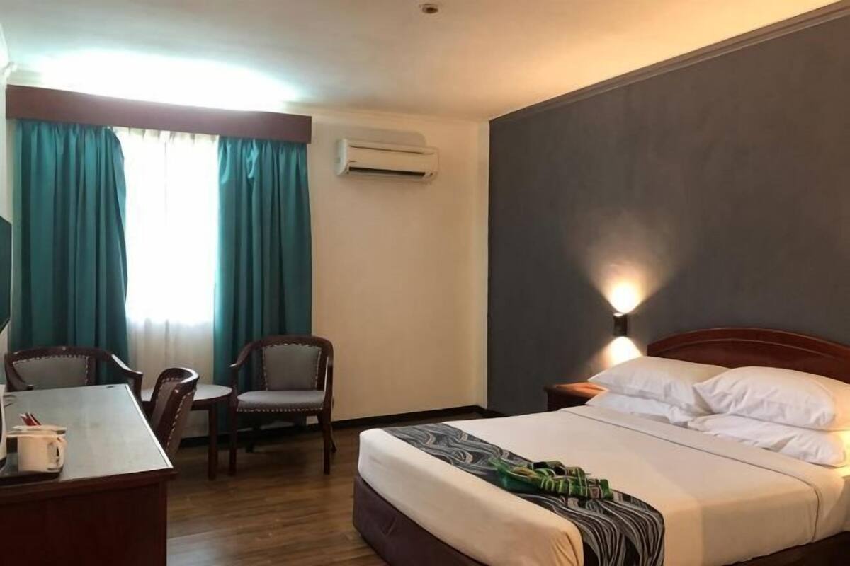 A hotel room with a bed, a desk, two chairs, a window with curtains, and a wall-mounted air conditioner.
