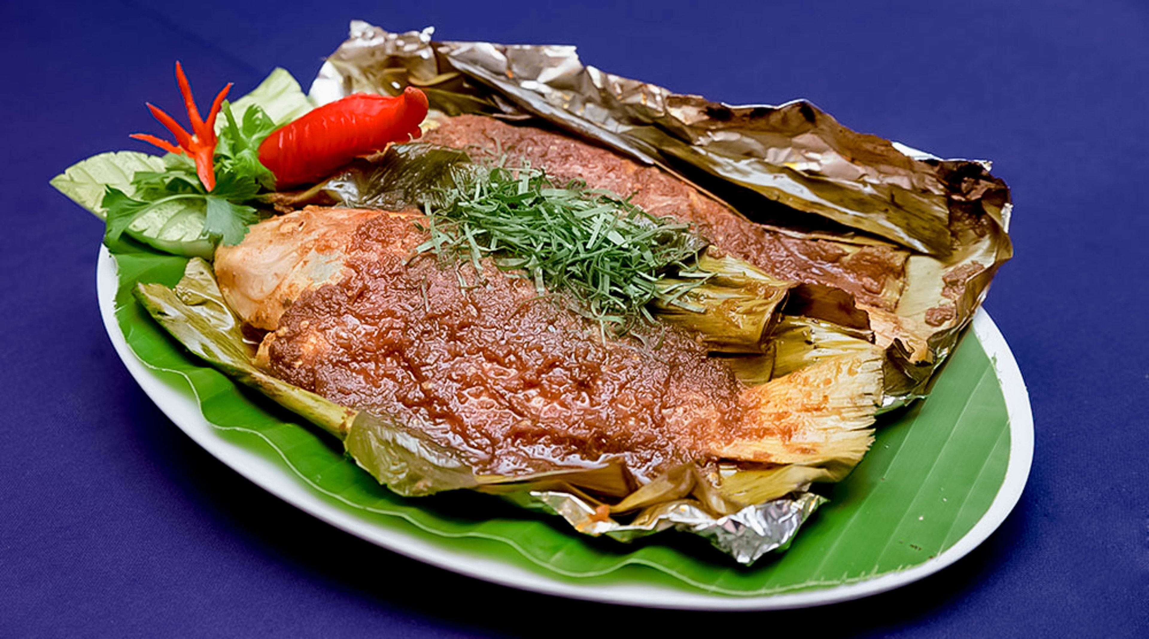 Pescado envuelto en hojas de plátano, adornado con hierbas y un chile, servido en un plato de hoja verde.