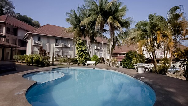 A swimming pool with a curved edge, surrounded by palm trees and a building with a red-tiled roof. - Hotel Seri Malaysia Temerloh (Temerloh)