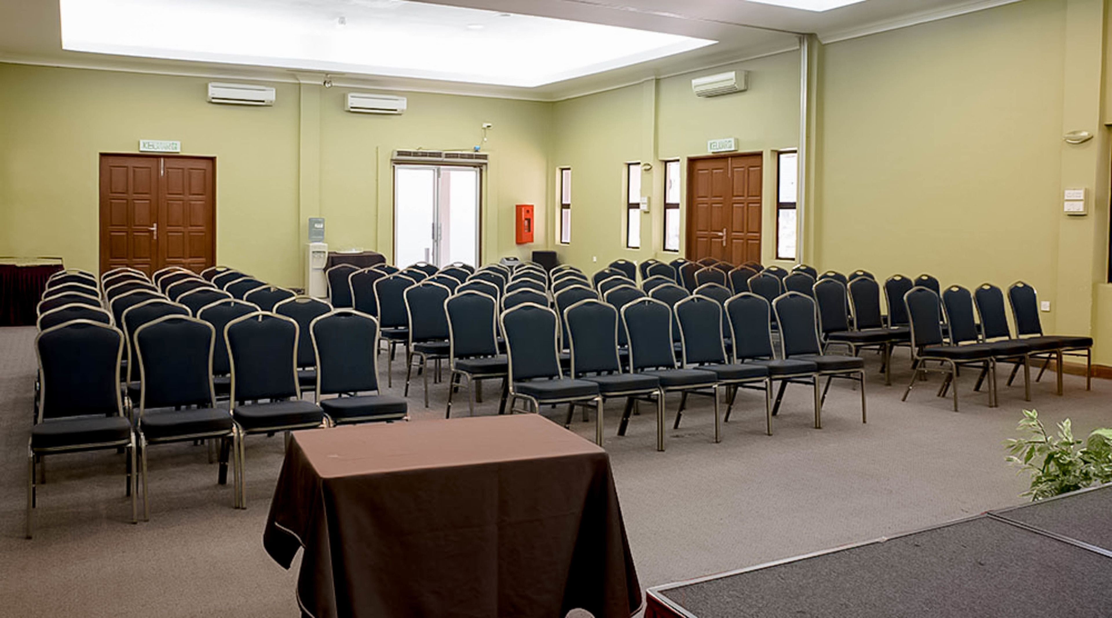 Un salón de conferencias con filas de sillas y mesas, unidades de aire acondicionado y puertas.
