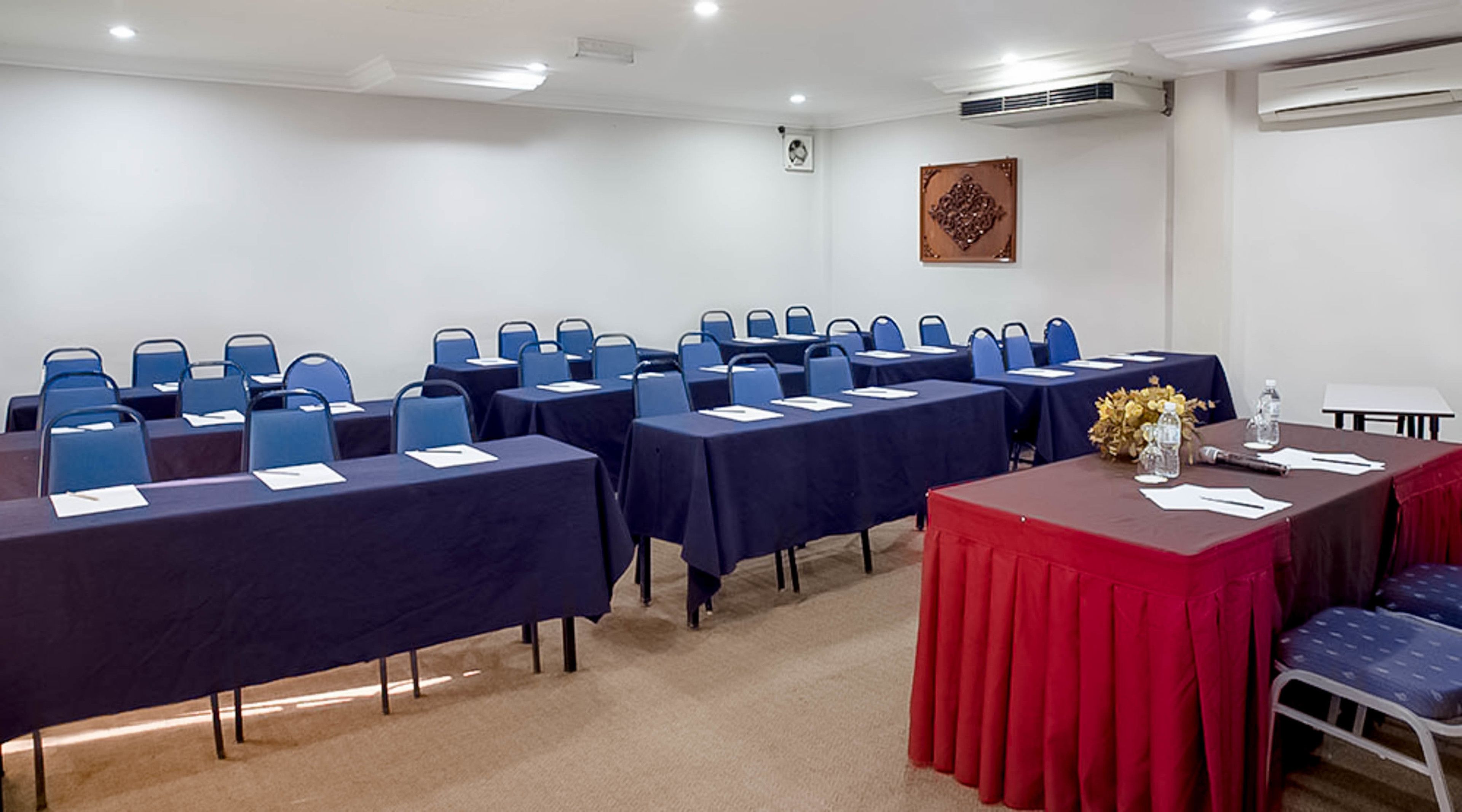 A conference room with rows of tables and chairs, a red table with a vase of flowers, and an air conditioning unit on the wall.