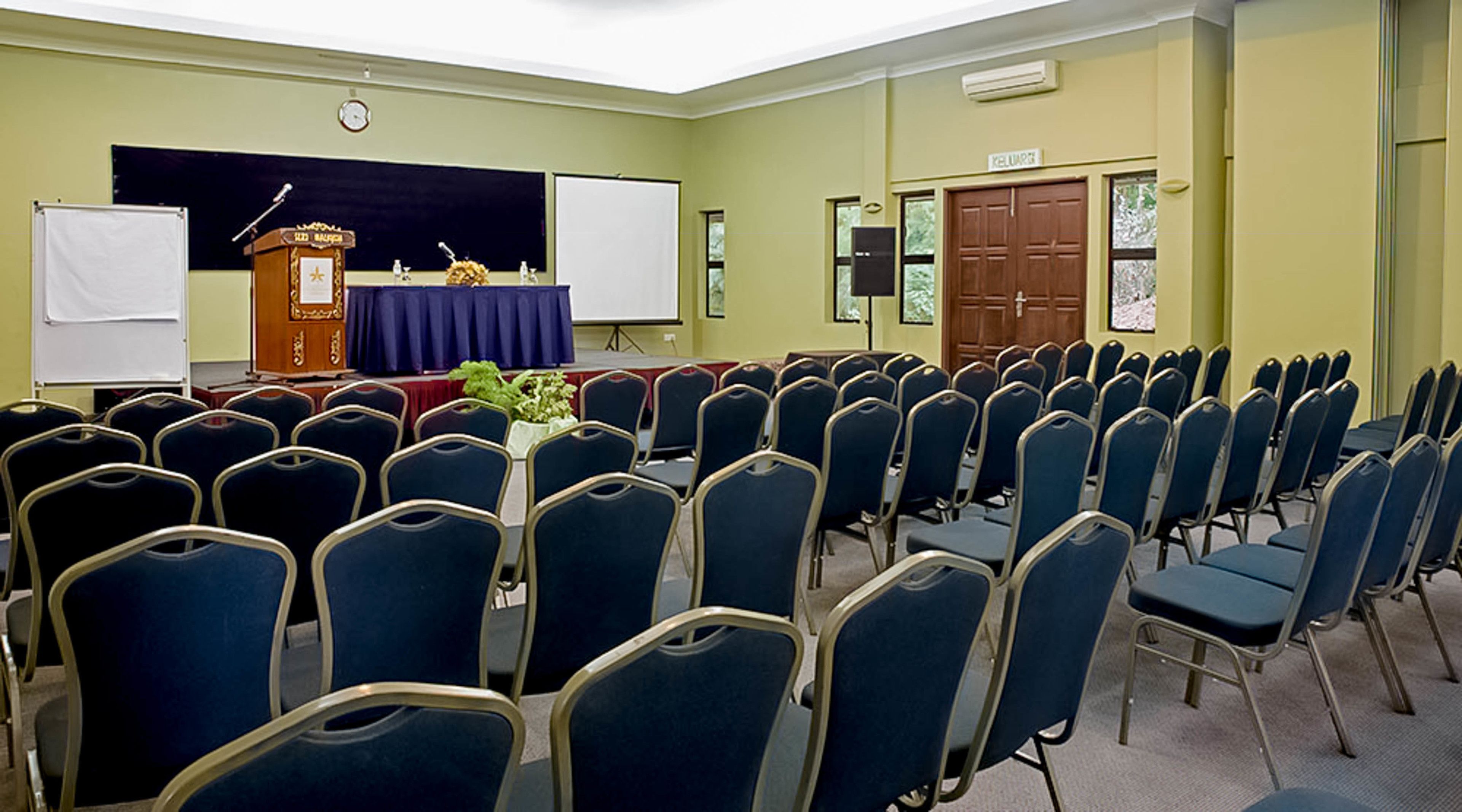 Un salón de conferencias con filas de sillas azules, un atril, una pizarra y una puerta con la etiqueta 'KELUAR'.