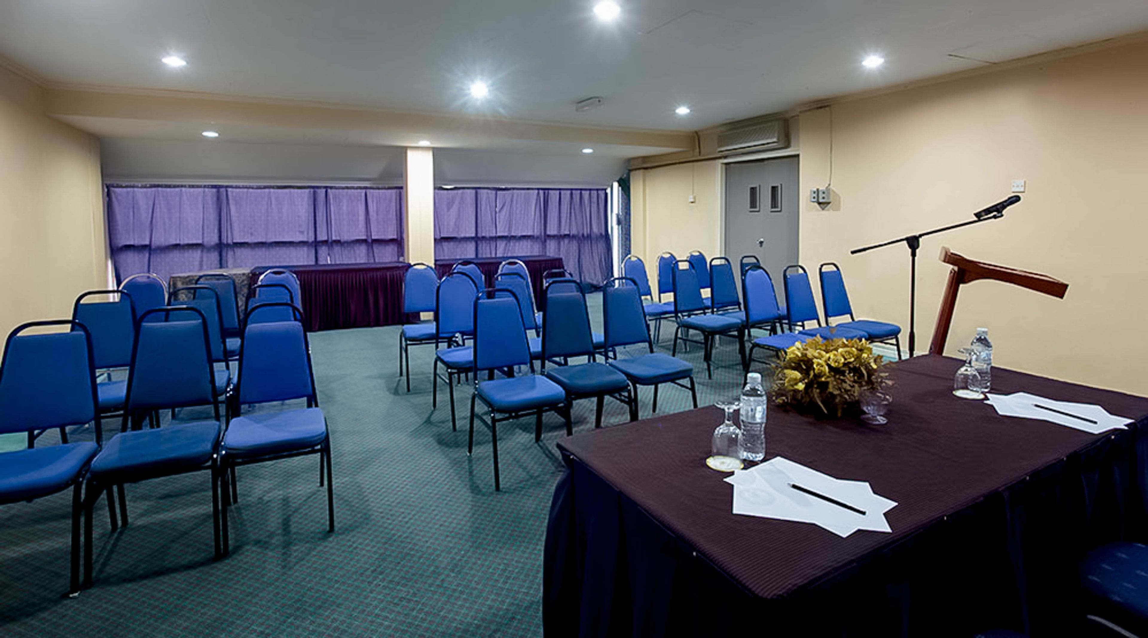 A conference room with rows of blue chairs, a table with water bottles and papers, and a microphone stand.