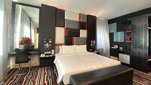 A modern hotel room with a large bed, a desk, a chair, a small table, a mirror, and a patterned carpet. - Swiss-Belcourt Makassar (Makassar)