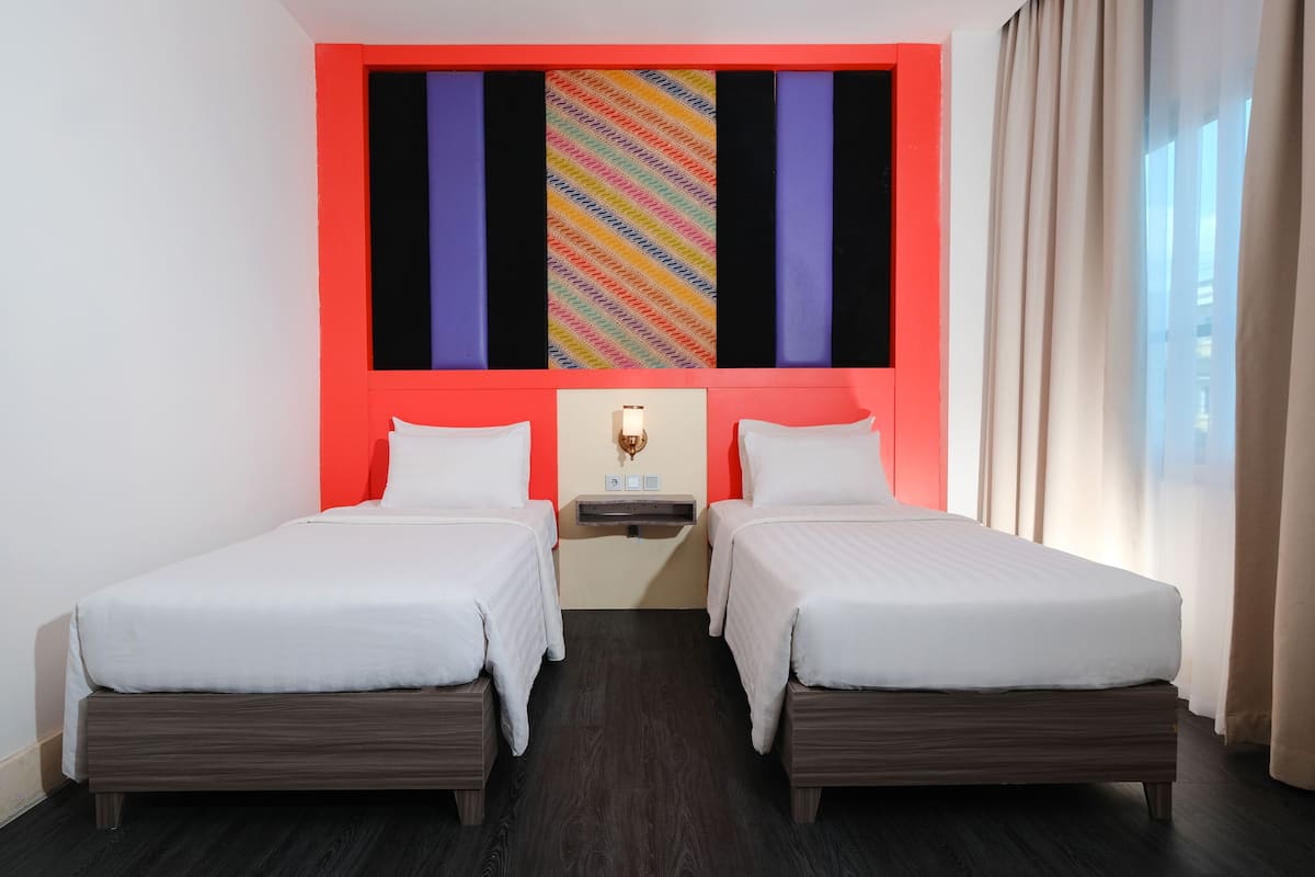 Habitación de hotel con dos camas individuales, una mesita de noche con lámpara, televisor montado en la pared y ventana con cortinas.