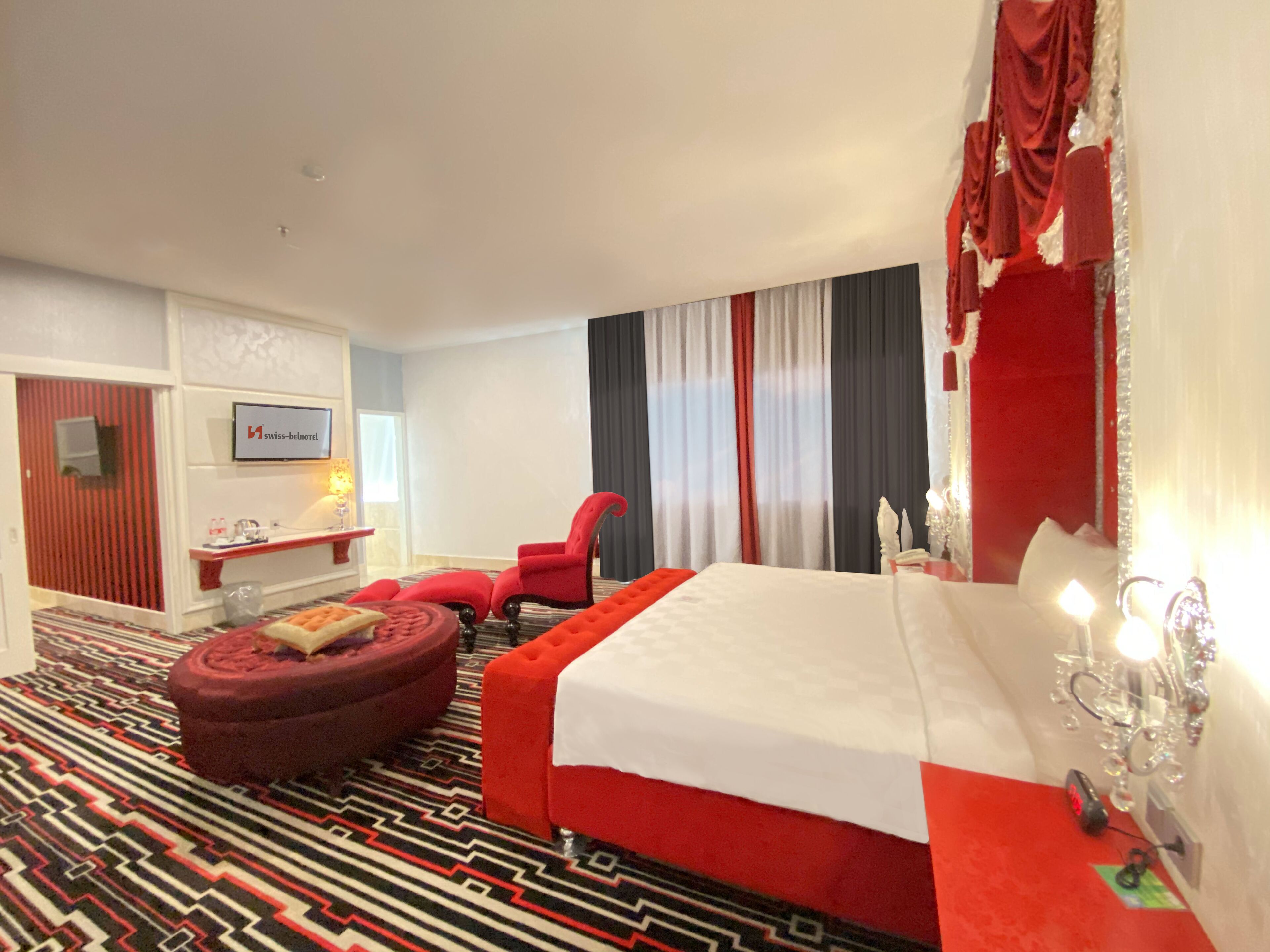A hotel room with a large bed, a red armchair, a small ottoman, a desk with a lamp, and a view of the city through the window.