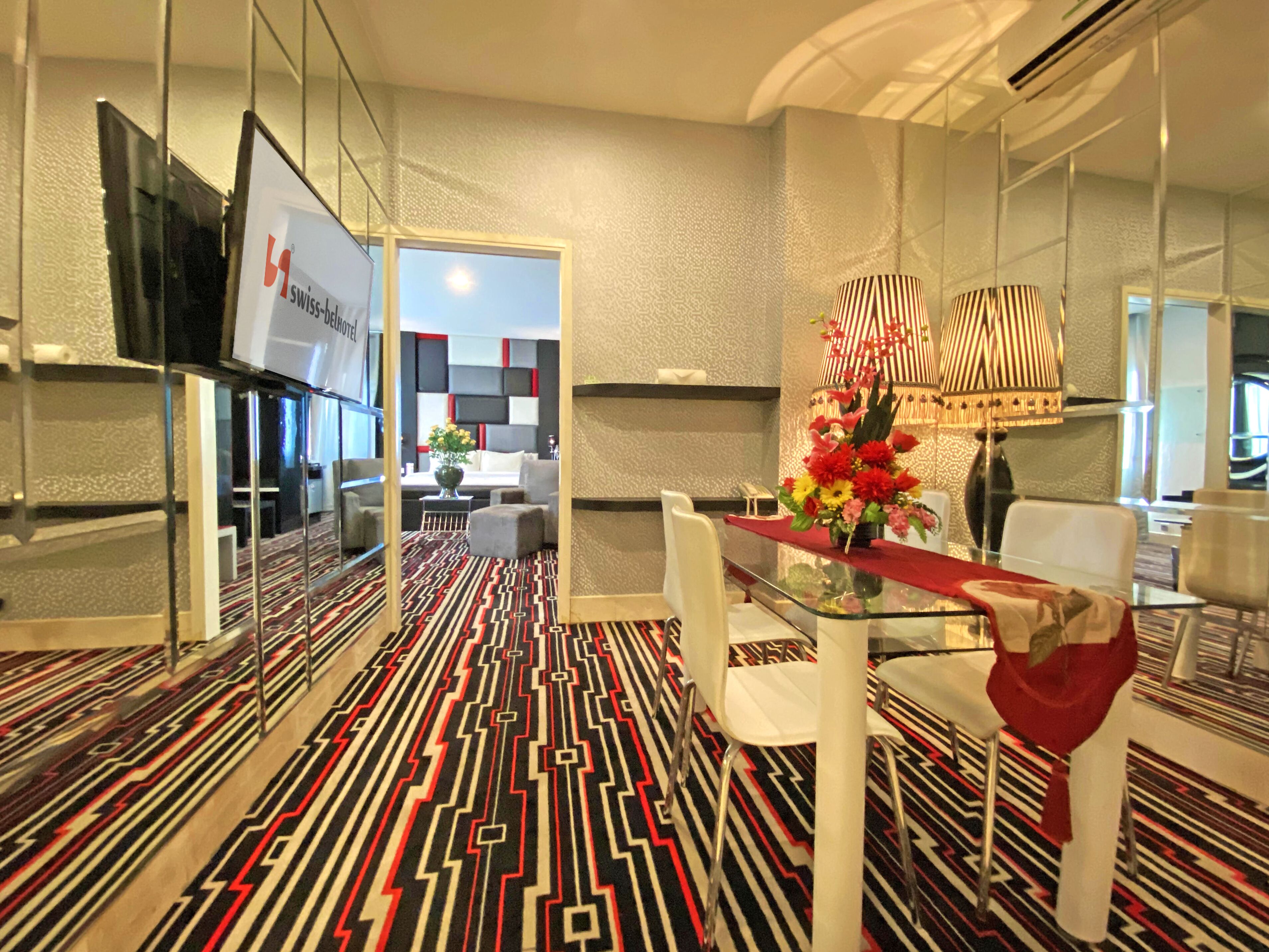 Un hall d’hôtel moderne avec un tapis à motifs, une table en verre entourée de chaises, une lampe décorative et un téléviseur fixé sur une cloison en verre.
