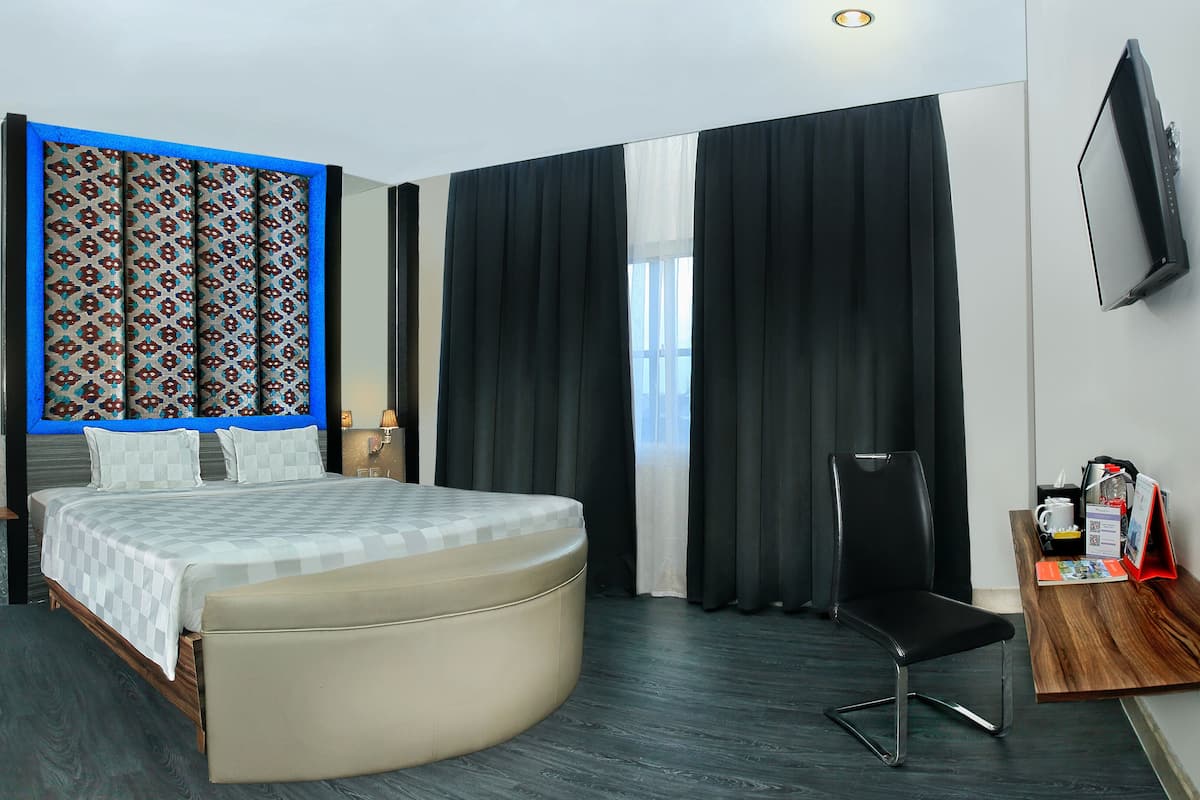 Habitación de hotel con cama, escritorio con silla, televisor montado en la pared y ventana con cortinas.