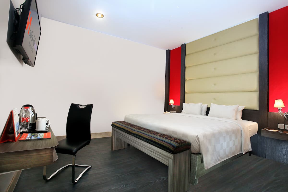 Una habitación de hotel con cama, escritorio con silla, televisor y mesita de noche con lámpara.