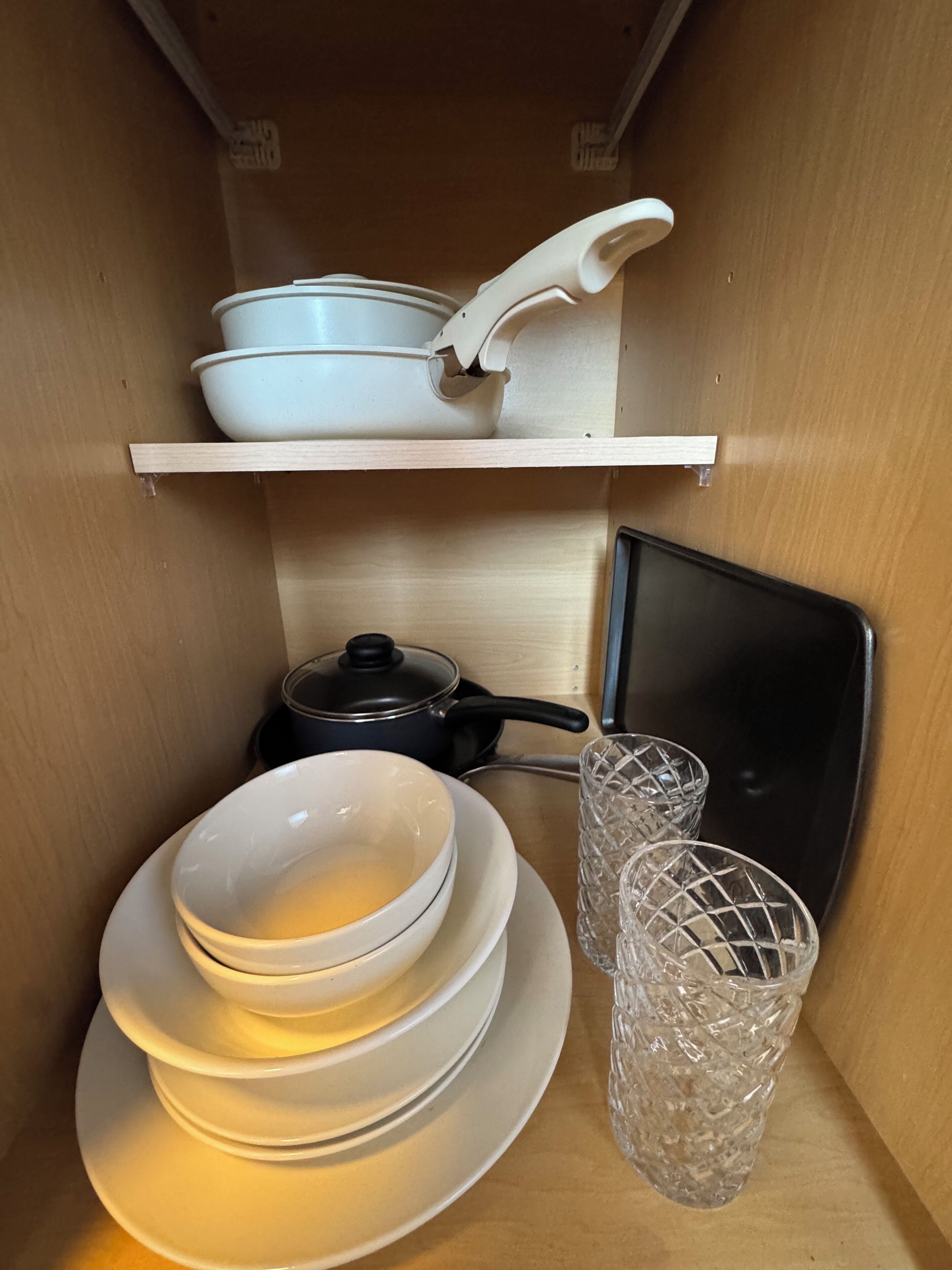 Een keuken met een witte plank waarop een witte pan en koekenpan staan, een zwarte pan op het aanrecht, en een glazen waterkan.