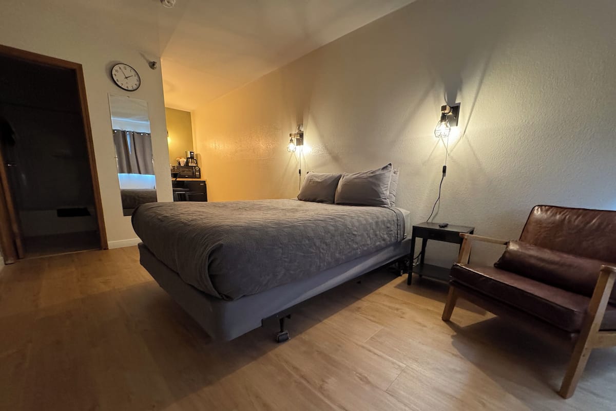 A bedroom with a bed, a chair, a nightstand, a clock, and a wall-mounted light fixture.
