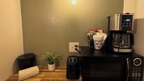 A corner of a room with a wooden floor, a black microwave, a toaster, a coffee maker, a small potted plant, and a wall-mounted light fixture. - The Pioneer Motel (Palmer)