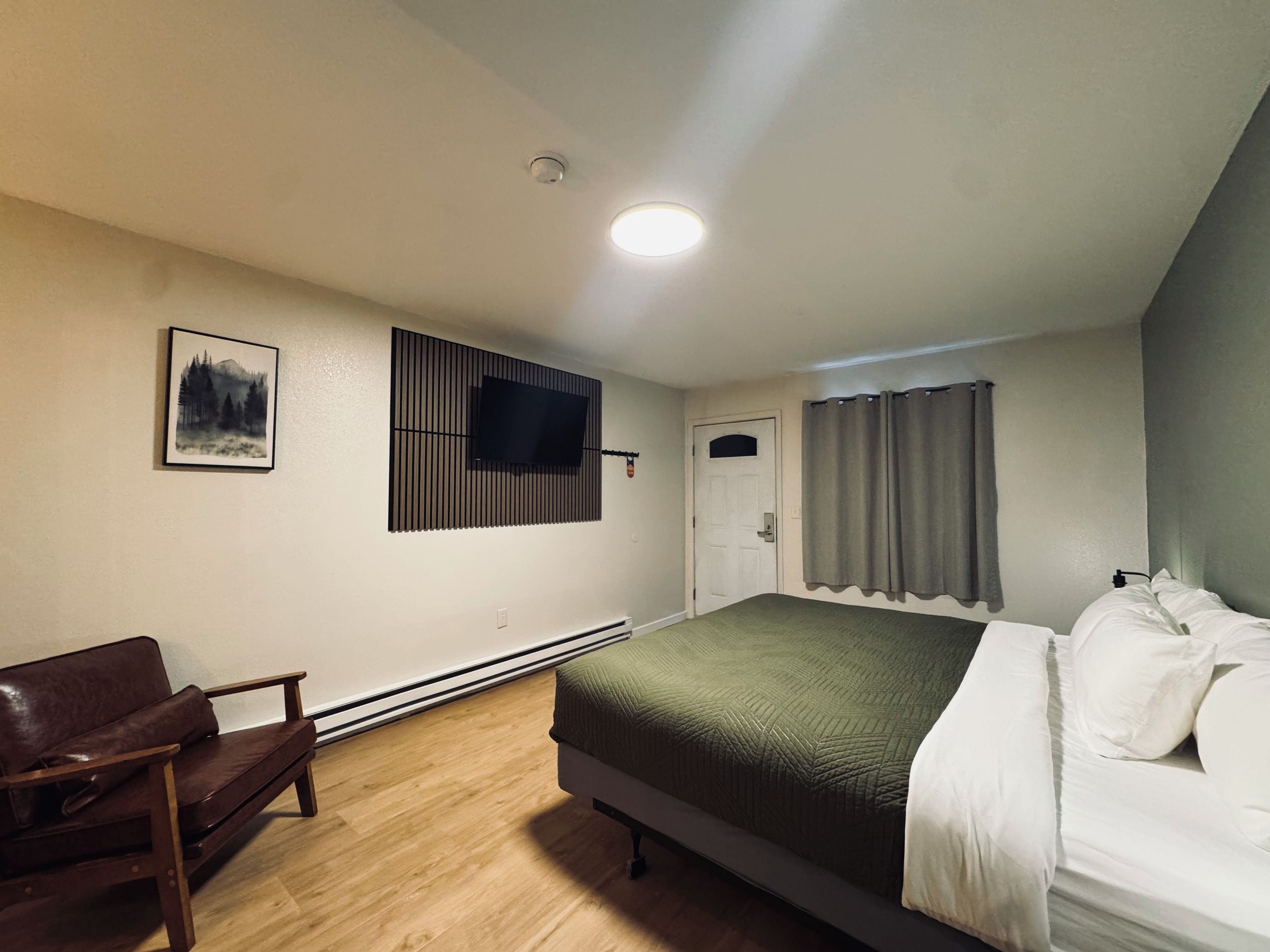 A bedroom with a bed, a chair, a TV mounted on the wall, a door, and a window with curtains.