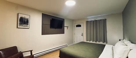 A bedroom with a bed, a chair, a TV mounted on the wall, a door, and a window with curtains.