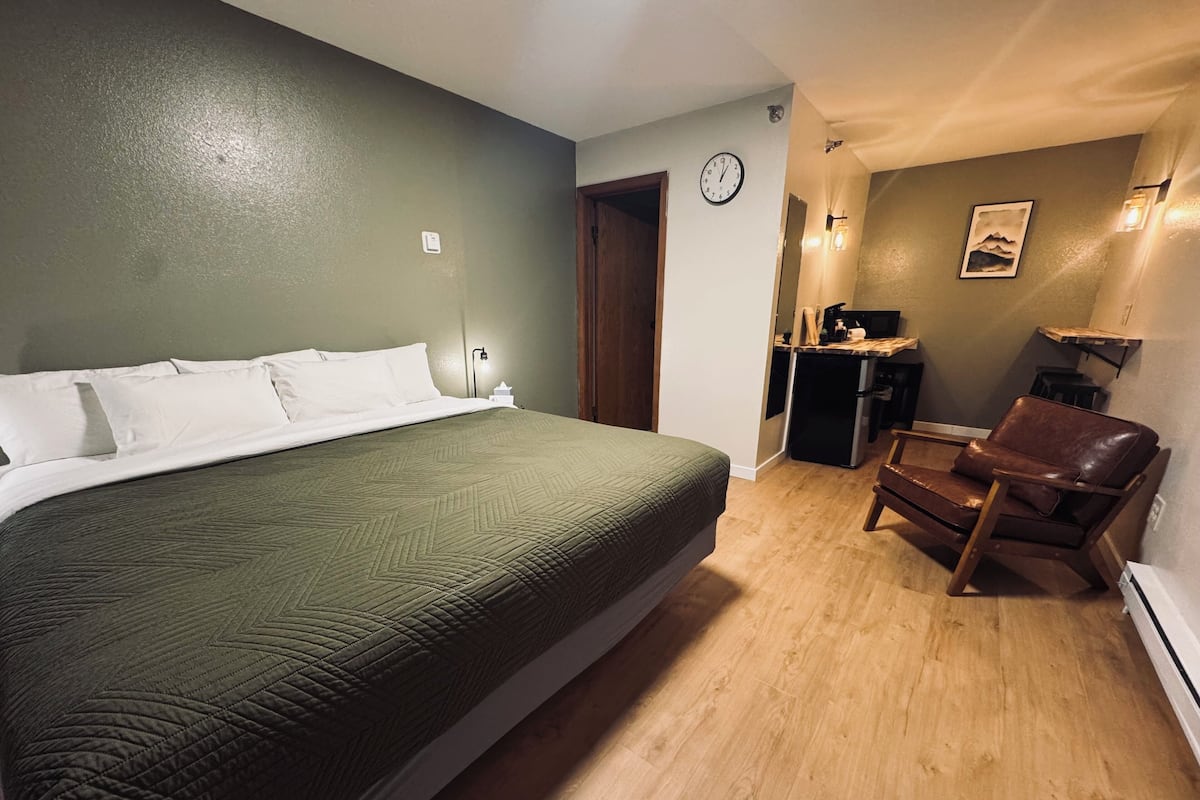 A bedroom with a large bed, a wooden chair, a small kitchen area, and a clock on the wall.