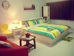 A bedroom with a bed, a nightstand, a chair, a small table with a lamp, and a clock on the wall. - Donmuang - Impact Condo Service (Pak Kret)