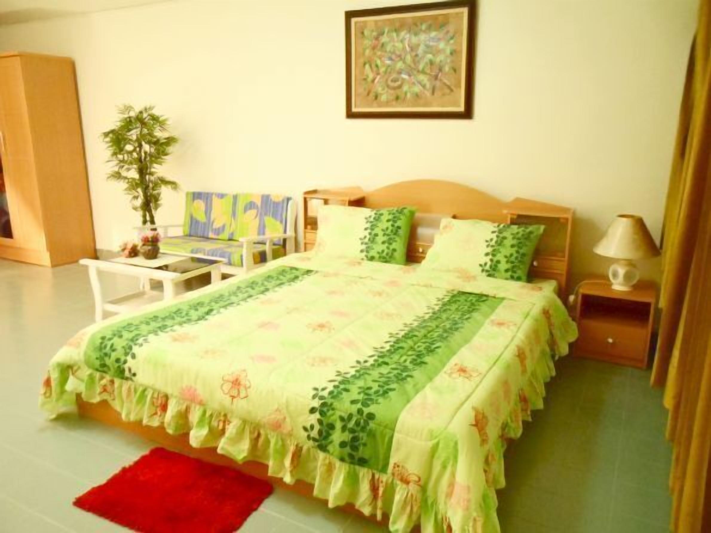 Un dormitorio con cama, mesita de noche, lámpara, silla, mesa, planta y un cuadro en la pared.