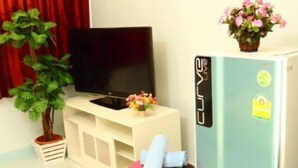 A compact living space with a flat-screen TV on a white cabinet, a small black chair, a pink ottoman, a refrigerator, and a potted plant. - Donmuang - Impact Condo Service (Pak Kret)