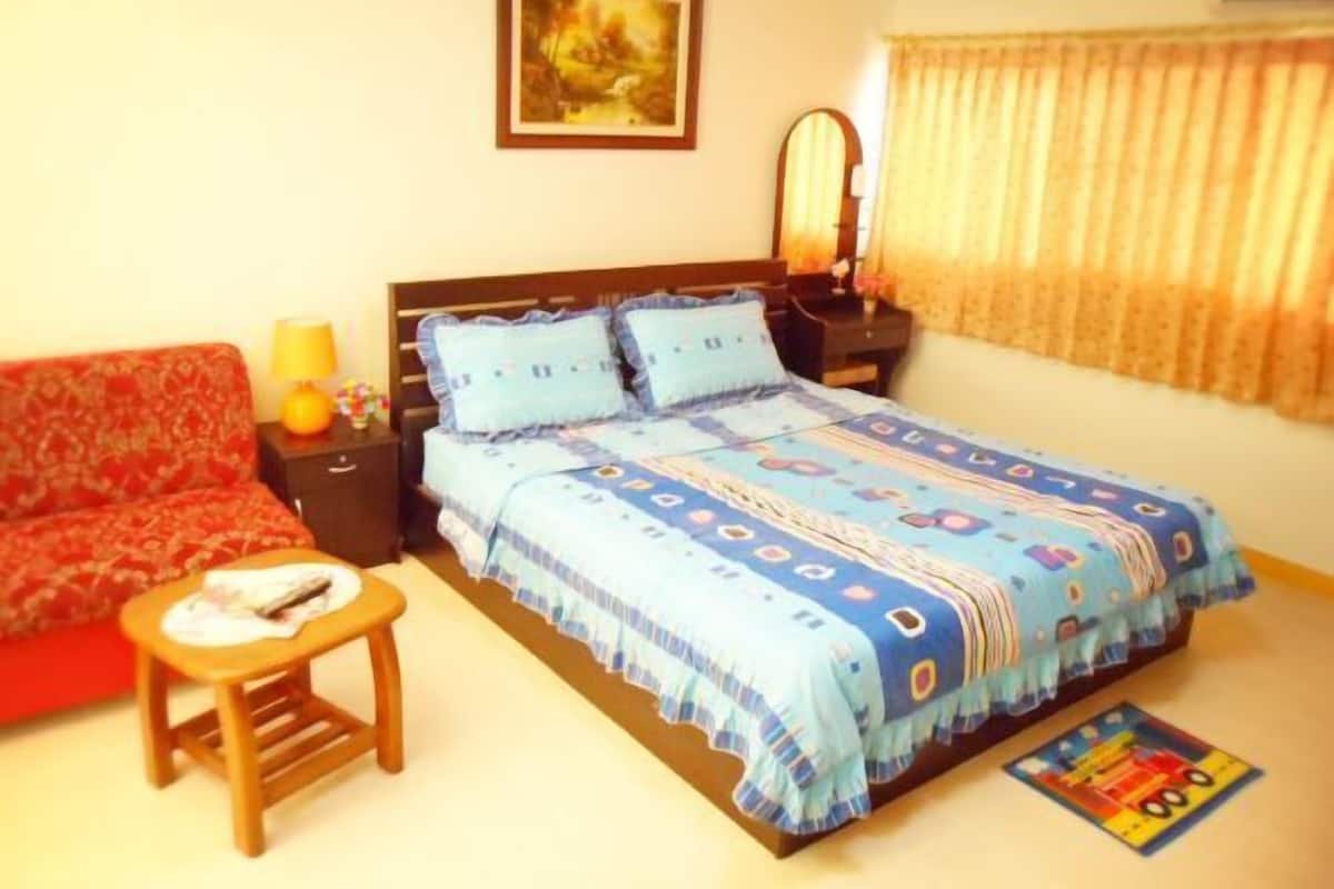 Quarto com cama, sofá vermelho, mesa de madeira pequena, mesa de cabeceira, luminária, espelho e janela com cortinas.